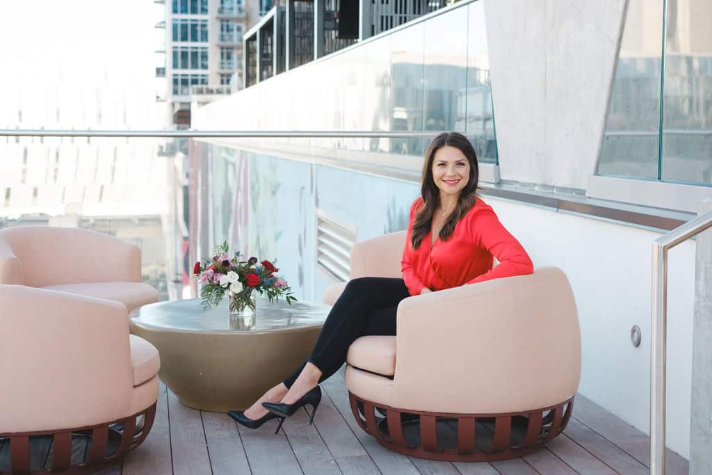Woman in red blouse sitting on pink outdoor lounge chair with flowers on table.
